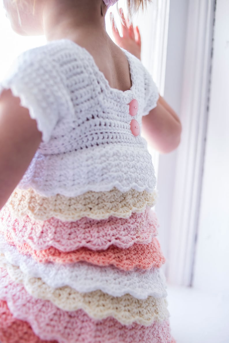 May include: A white crocheted dress with a layered skirt in shades of pink, peach, and cream. The dress has a scalloped hem and two pink buttons on the back.