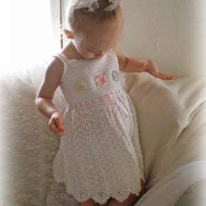 Könnte beinhalten: Ein weißes Häkelkleid mit einem gewellten Saum. Das Kleid hat ein ärmelloses Design mit kleinen floralen Verzierungen und einem rosa Band. Das Kleid ist für ein Kind entworfen.