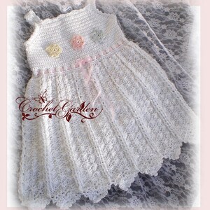 May include: A white crochet dress with a floral pattern and scalloped hem. The dress features a bodice with three pastel-colored crochet flowers and a pink ribbon. The text "Crochet Garden" is visible on the lower left.