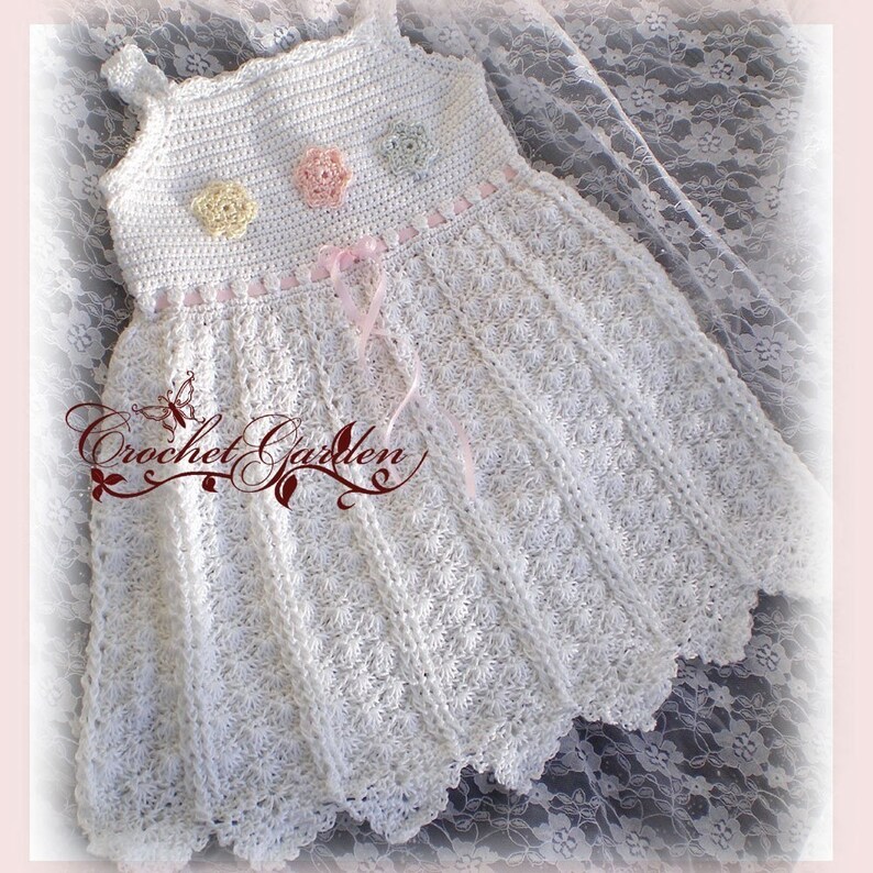 May include: A white crochet dress with a floral pattern and scalloped hem. The dress features a bodice with three pastel-colored crochet flowers and a pink ribbon. The text "Crochet Garden" is visible on the lower left.