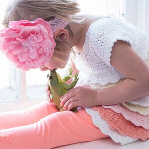 May include: A young girl wearing a pink flower headband and a white crocheted dress with layers of pink and peach ruffles is kissing a green frog toy.