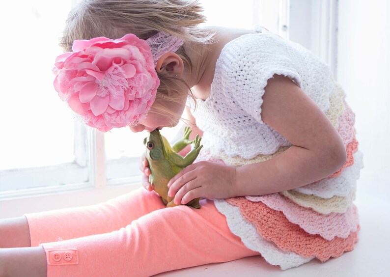 May include: A young girl wearing a pink flower headband and a white crocheted dress with layers of pink and peach ruffles is kissing a green frog toy.