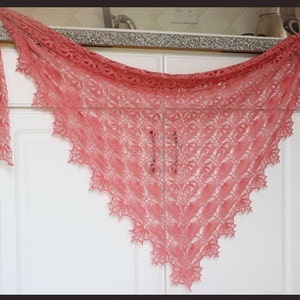 Puede incluir: Un chal de encaje delicado de color coral con un borde festoneado. El chal está drapeado sobre una encimera blanca.