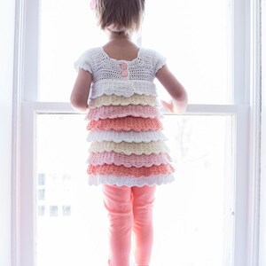 May include: A young girl wearing a white crocheted dress with pink, peach, and yellow ruffles. The dress has two pink buttons on the back. The girl is standing in front of a window and looking out.