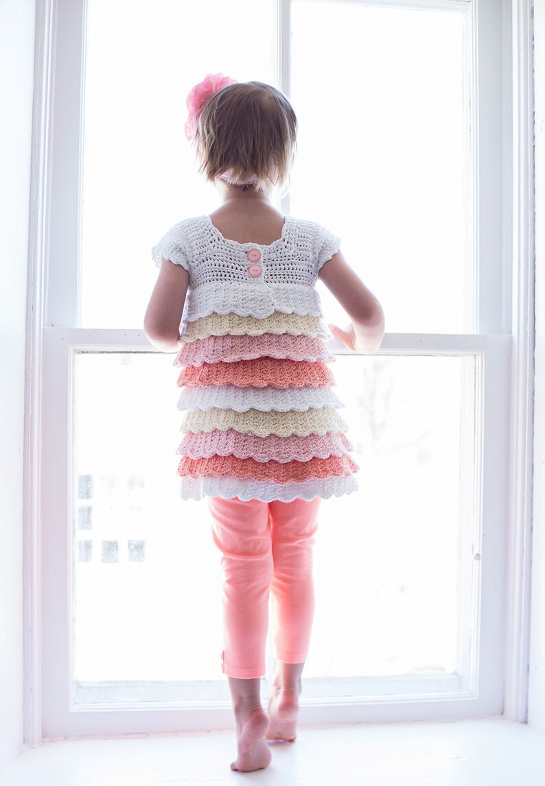 May include: A young girl wearing a white crocheted dress with pink, peach, and yellow ruffles. The dress has two pink buttons on the back. The girl is standing in front of a window and looking out.