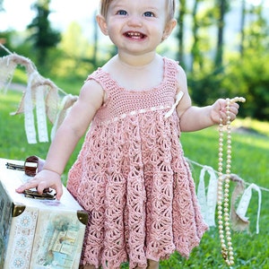May include: A young girl wearing a pink crocheted dress and a flower headband smiles while holding a pearl necklace. She is standing in a grassy area with a vintage suitcase beside her.