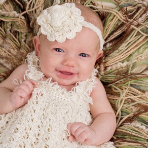 May include: A baby wearing a white crocheted dress and a white crocheted flower headband. The baby is lying on a brown and green textured surface.