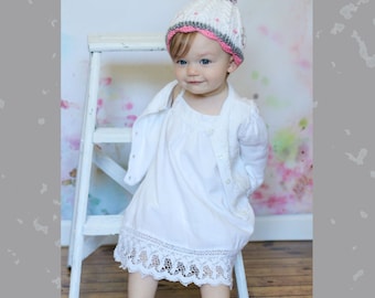 May include: A young child wearing a white crocheted hat with pink and gray accents and a white dress with lace trim. The child is sitting on a white wooden step stool.