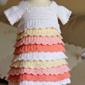 May include: A white crocheted baby dress with a scalloped hem. The dress has multiple layers of ruffles in shades of peach, pink, and yellow.