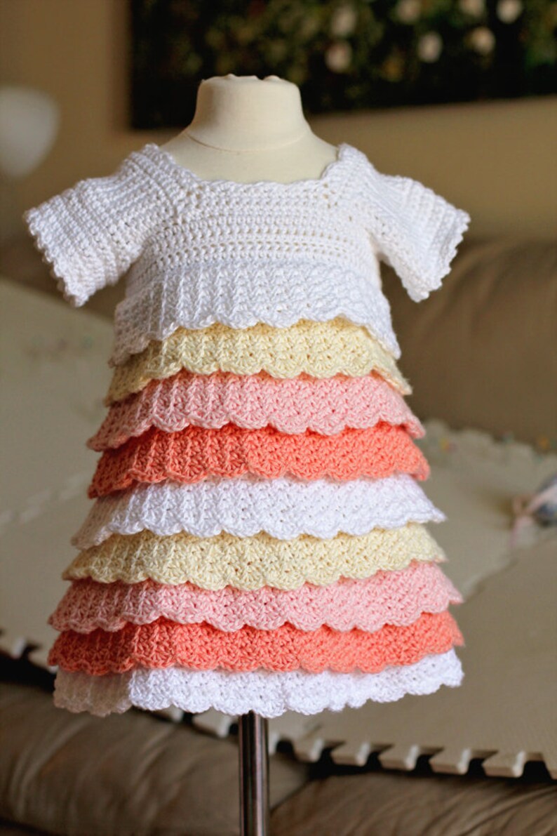 May include: A white crocheted baby dress with a scalloped hem. The dress has multiple layers of ruffles in shades of peach, pink, and yellow.