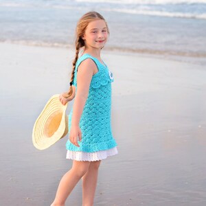 K&ouml;nnte beinhalten: Ein junges M&auml;dchen in einem blauen H&auml;kelkleid mit wei&szlig;er Spitzenborte steht an einem Sandstrand. Sie h&auml;lt einen Strohhut in der Hand und l&auml;chelt in die Kamera.