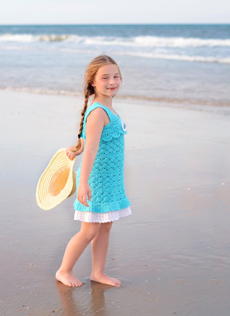 K&ouml;nnte beinhalten: Ein junges M&auml;dchen in einem blauen H&auml;kelkleid mit wei&szlig;er Spitzenborte steht an einem Sandstrand. Sie h&auml;lt einen Strohhut in der Hand und l&auml;chelt in die Kamera.