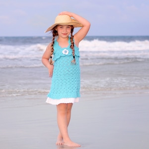 K&ouml;nnte beinhalten: Ein junges M&auml;dchen in einem hellblauen H&auml;kelkleid mit wei&szlig;em Spitzenbesatz und einem Strohhut steht an einem Sandstrand. Im Hintergrund sind die Wellen des Ozeans zu sehen.