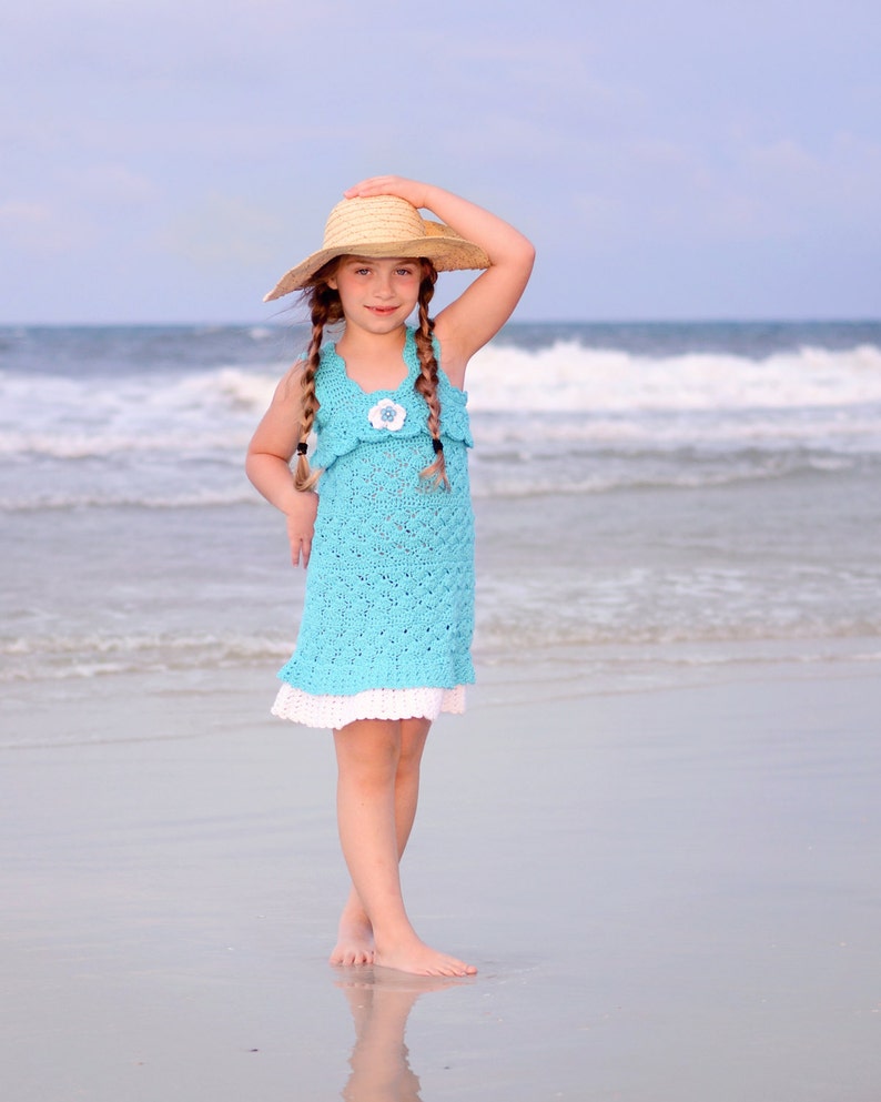 K&ouml;nnte beinhalten: Ein junges M&auml;dchen in einem hellblauen H&auml;kelkleid mit wei&szlig;em Spitzenbesatz und einem Strohhut steht an einem Sandstrand. Im Hintergrund sind die Wellen des Ozeans zu sehen.