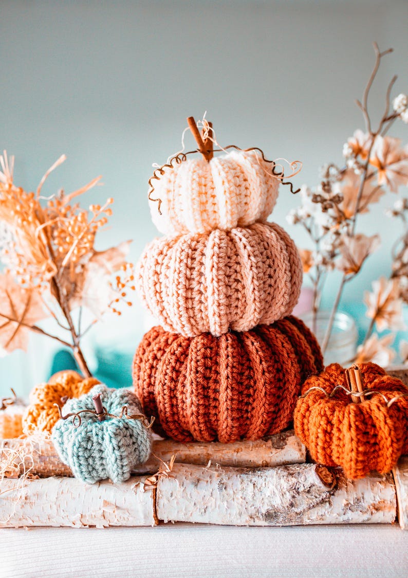 May include: A stack of three crocheted pumpkins in shades of cream, peach, and rust-orange, with a small, teal pumpkin in front. The pumpkins are arranged on a birch log, with dried flowers in the background. The pumpkins have cinnamon stick stems.