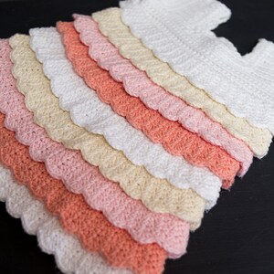 May include: A crocheted baby dress with a scalloped edge. The dress is made in shades of white, cream, pink, and coral.