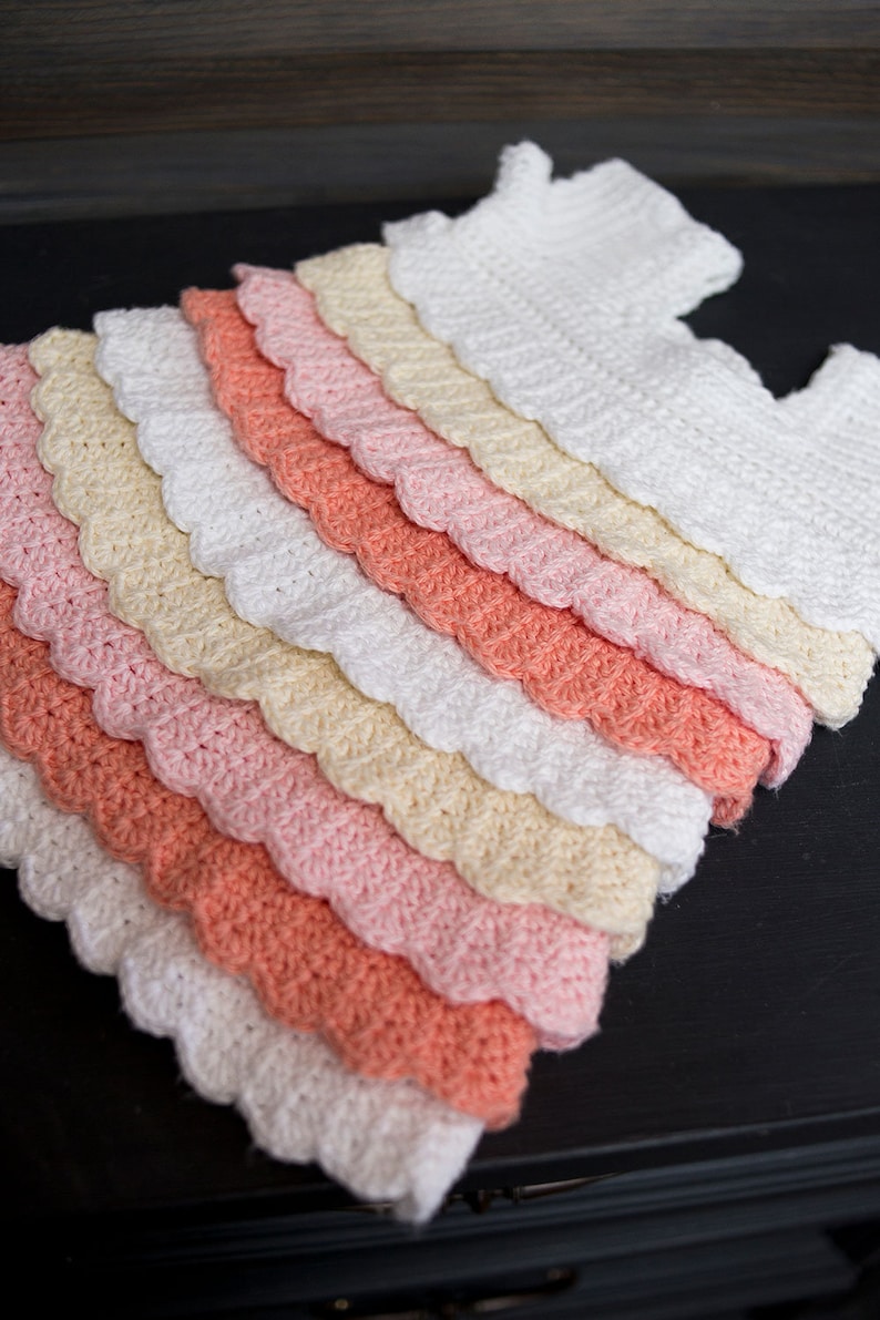 May include: A crocheted baby dress with a scalloped edge. The dress is made in shades of white, cream, pink, and coral.