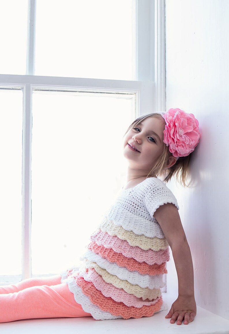 May include: A young girl wearing a white crocheted top with multiple layers of pink, peach, and yellow ruffles. She is wearing a pink flower headband and has a bright smile.