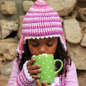 May include: A child wearing a pink, white, and purple crocheted hat and matching mittens holds a green mug with white polka dots. The mug is filled with marshmallows. The child is wearing a pink long-sleeved shirt.