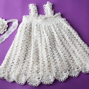 May include: A white crocheted baby dress with a matching headband. The dress has a scalloped hem and a delicate floral design. The headband features a white flower.