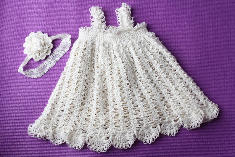 May include: A white crocheted baby dress with a matching headband. The dress has a scalloped hem and a delicate floral design. The headband features a white flower.