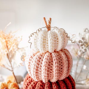 May include: A stack of three crocheted pumpkins in shades of cream, peach, and rust. The top pumpkin has a cinnamon stick stem and decorative wire tendrils. The pumpkins are a fall-themed decorative item.