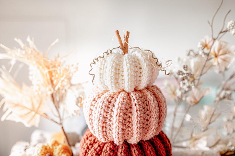 May include: A stack of three crocheted pumpkins in shades of cream, peach, and rust. The top pumpkin has a cinnamon stick stem and decorative wire tendrils. The pumpkins are a fall-themed decorative item.