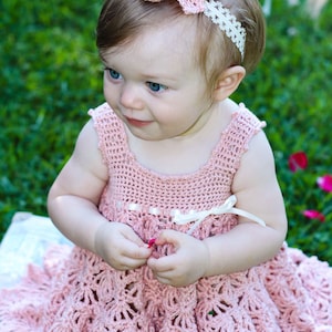 May include: A baby girl wearing a pink crocheted dress with a white crocheted flower headband. The dress has a scalloped hem and a white ribbon around the waist.