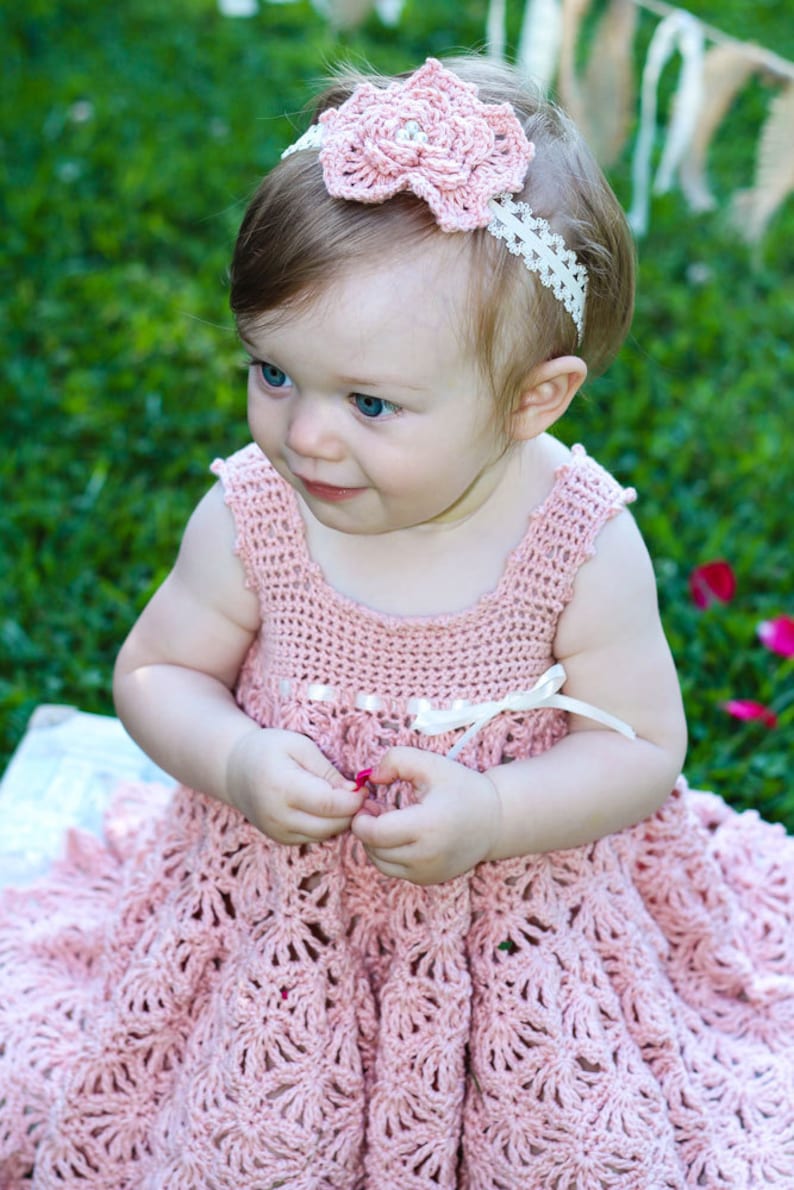 May include: A baby girl wearing a pink crocheted dress with a white crocheted flower headband. The dress has a scalloped hem and a white ribbon around the waist.