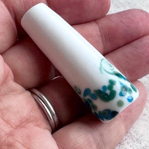 May include: A cylindrical white ceramic bead with a tapered shape, featuring a decorative pattern of small, scattered spots in shades of teal, dark green, and light blue concentrated toward the wider end of the piece.