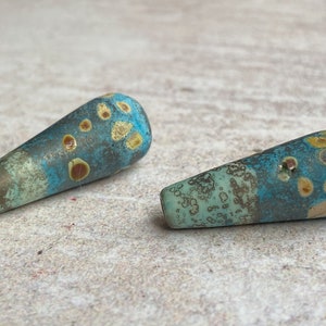 May include: Two teal blue and brown glass beads with a speckled pattern. The beads are shaped like a teardrop and have a glossy finish.