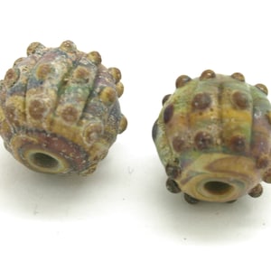 May include: Two handmade glass beads with a textured, organic design. The beads feature a mix of colours, including brown, yellow, purple, and green. Each bead has a central hole and is adorned with small, raised dots. These beads are suitable for jewellery making.