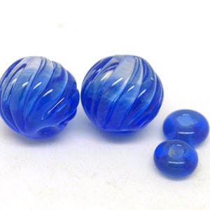 May include: A selection of blue glass beads. Two large, round beads with a spiral design sit beside three smaller, oval-shaped beads. The beads are a vibrant shade of blue and appear translucent.