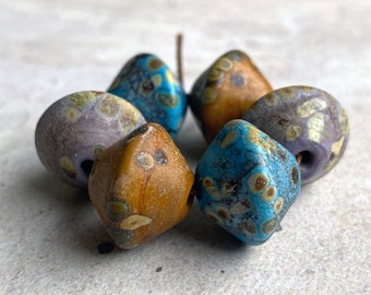 May include: A group of six ceramic beads with a speckled glaze in shades of blue, purple, and brown. The beads are shaped like irregular pyramids.