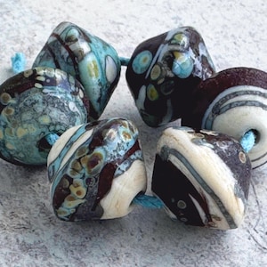 May include: A collection of five handcrafted glass beads strung on a light blue cord. The beads feature a mix of dark brown, cream, and turquoise blue colours with swirling patterns, speckled details, and smooth, rounded, or slightly faceted shapes.