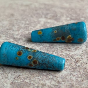 May include: Two turquoise blue glass beads with brown and yellow flecks. The beads are cone shaped and have a glossy finish.