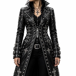 May include: A black long-length coat featuring silver-toned metal studs, multiple adjustable buckle straps along the waist and sleeves, and embroidered cross motifs. The garment includes a high collar, a central zip closure, and a structured fit.