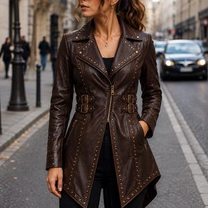 May include: A dark brown leather jacket featuring a high-low hemline, gold-toned metal stud embellishments along the lapels and front panels, a central zipper closure, and two adjustable buckled straps at the waist with matching gold hardware.