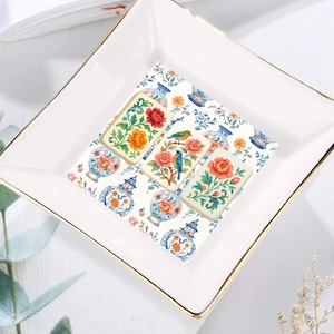 May include: A square, white ceramic trinket dish with gold trim. The dish holds three decorative tiles with floral and bird designs in red, orange, and blue. The tiles sit on a patterned background of vases and flowers.