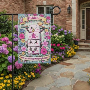 Puede incluir: Una bandera de jardín con una pila de fichas de mahjong de dibujos animados con caras y caracteres chinos. La bandera tiene el texto "Shhhh..." y "We are playing Mahjong inside!" rodeado de flores. Se muestra en un soporte de metal negro.