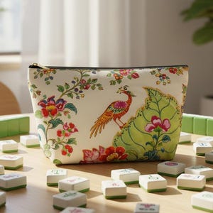 Może przedstawiać: Kremowa kosmetyczka zapinana na zamek z kwiatowym i ptasim wzorem. Kosmetyczka przedstawia kolorowego ptaka siedzącego na dużym zielonym liściu, otoczonego czerwonymi i różowymi kwiatami. Kosmetyczka leży na drewnianym stole z kafelkami do gry w Mahjonga.
