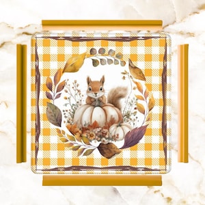 May include: A square decorative tray with a fall theme. The tray features a watercolor illustration of a squirrel sitting on a pumpkin, surrounded by leaves and flowers. The background has a yellow and white checkered pattern.