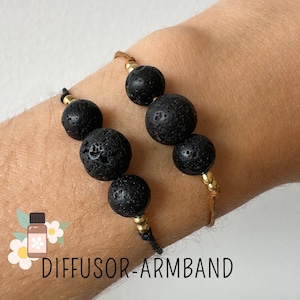 Könnte beinhalten: Zwei schwarze Lava-Diffusor-Armbänder mit goldenen Perlen und braunem Band. Die Armbänder werden am Handgelenk einer Person getragen. Der Text "DIFFUSOR-ARMBAND" ist unterhalb der Armbänder sichtbar.