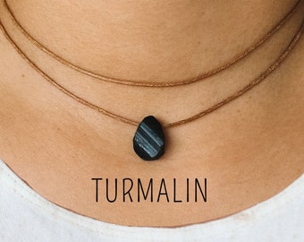 May include: A black tourmaline gemstone pendant on a brown cord necklace. The necklace is layered and the pendant is a teardrop shape. The text "TURMALIN" is written below the pendant.