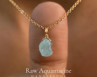 May include: A gold chain necklace featuring a raw, light blue aquamarine pendant wrapped in gold wire. The text "Raw Aquamarine HANDFREUDE" is printed in white at the bottom of the image, centered below the stone pendant.
