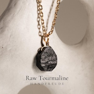 Könnte beinhalten: Eine Halskette mit einem Anhänger aus rohem schwarzem Turmalin, der an einer goldfarbenen Metallkette hängt. Der Anhänger ist mit goldfarbenem Draht umwickelt. Der Text "Raw Tourmaline" und "HANDFREUDE" steht unten im Bild.