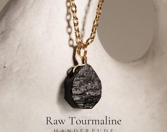 Puede incluir: Un collar con un colgante de turmalina negra en bruto suspendido de una cadena de metal de color dorado. El colgante está sujeto con un alambre de color dorado. El texto "Raw Tourmaline" y "HANDFREUDE" aparece en la parte inferior.