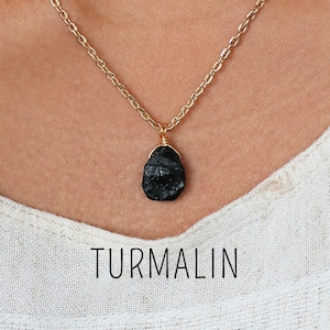 Puede incluir: Un collar de cadena dorada con un colgante de turmalina negra en bruto. El colgante está envuelto en alambre y cuelga de la cadena. La palabra "TURMALIN" está impresa en negro, en una fuente sans-serif, debajo del collar.