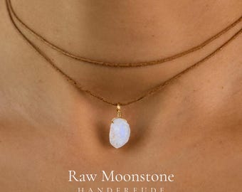 May include: A double-strand brown cord necklace featuring a single raw moonstone pendant wrapped in gold-toned wire. The text Raw Moonstone HANDFREUDE is printed in white at the bottom of the image.