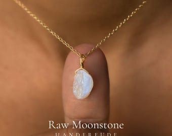 May include: A gold chain necklace featuring a raw, irregular moonstone pendant wrapped in gold wire. The stone displays a translucent, milky white appearance with subtle blue iridescence. The text Raw Moonstone HANDFREUDE is visible.